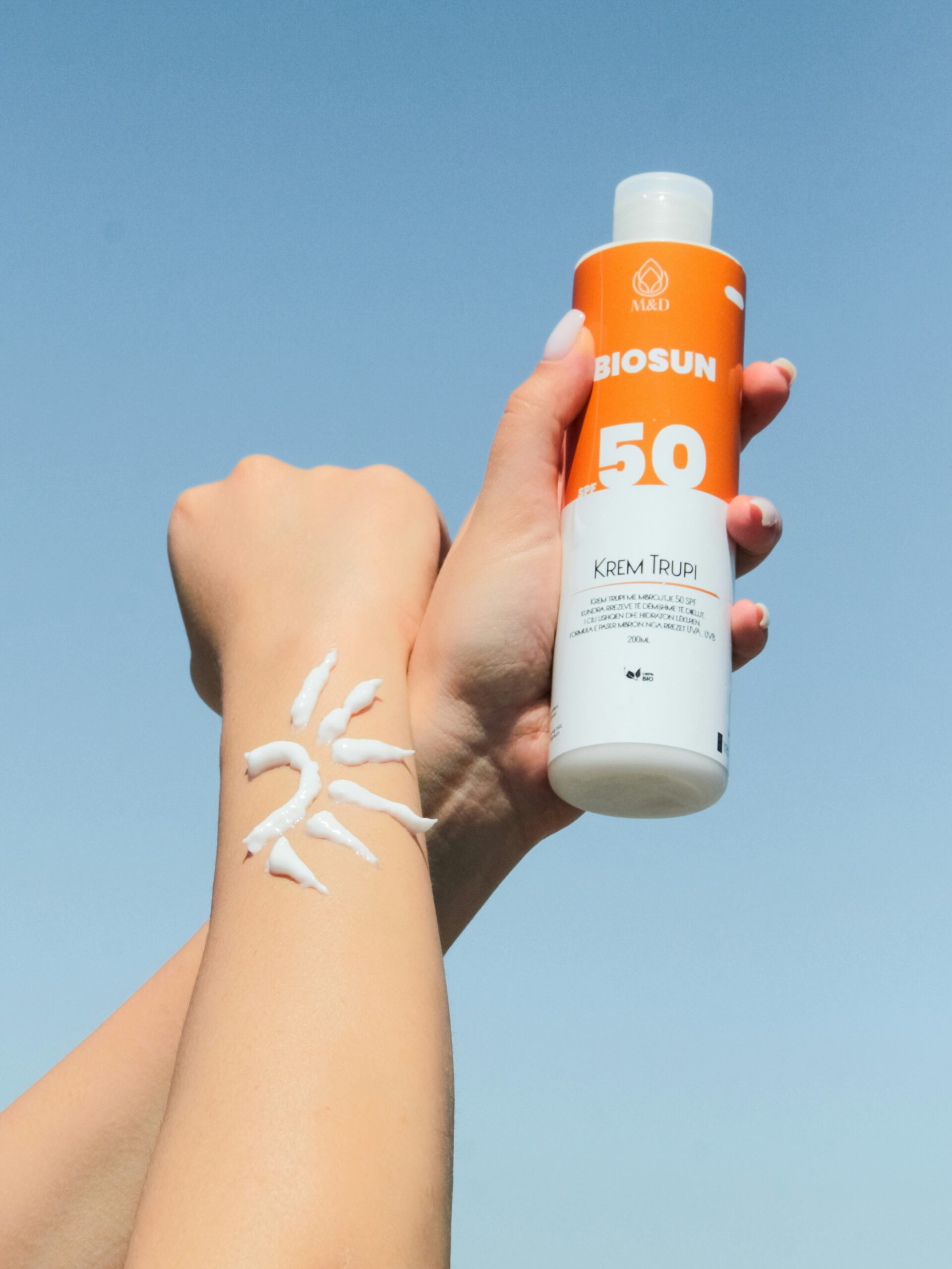 Gros plan sur les bras d’une femme sur fond de ciel bleu : une main tient un flacon de crème solaire, l’autre avant-bras porte de la crème solaire appliquée en forme de soleil