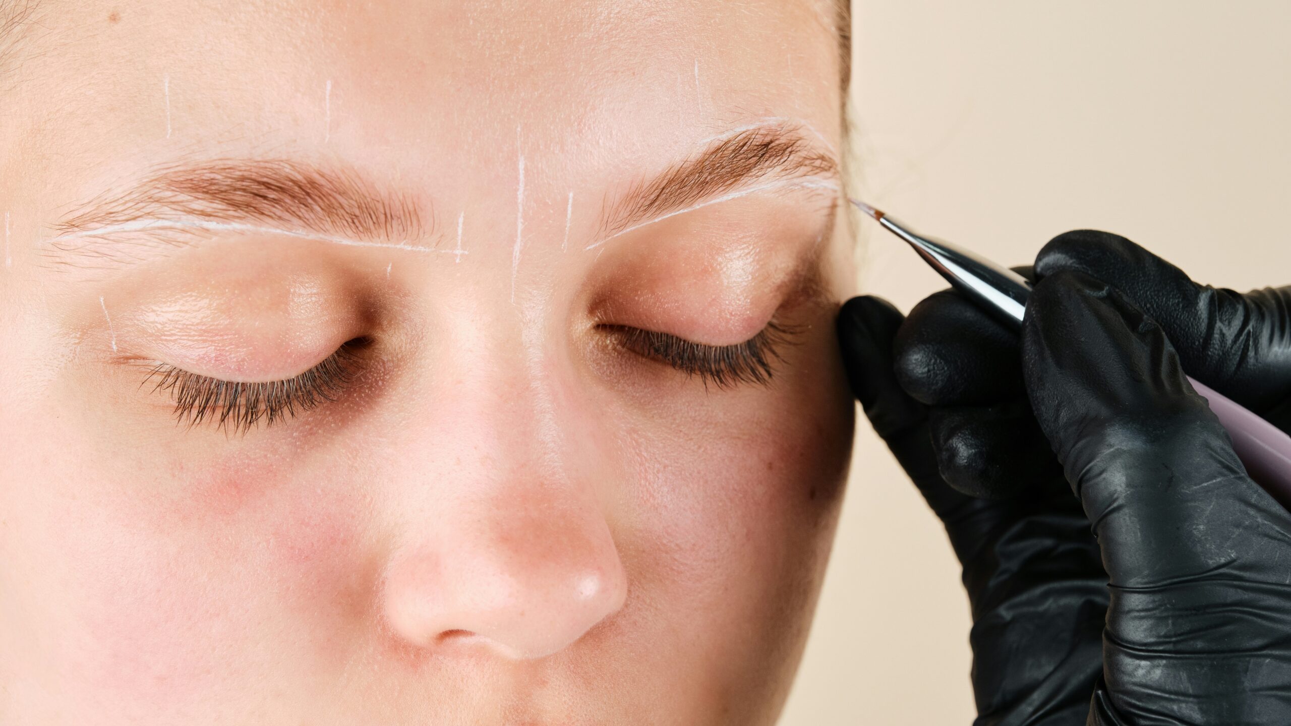 Gros plan sur le visage d’une femme pendant qu’une technicienne dessine des repères blancs pour tracer la forme souhaitée de ses sourcils