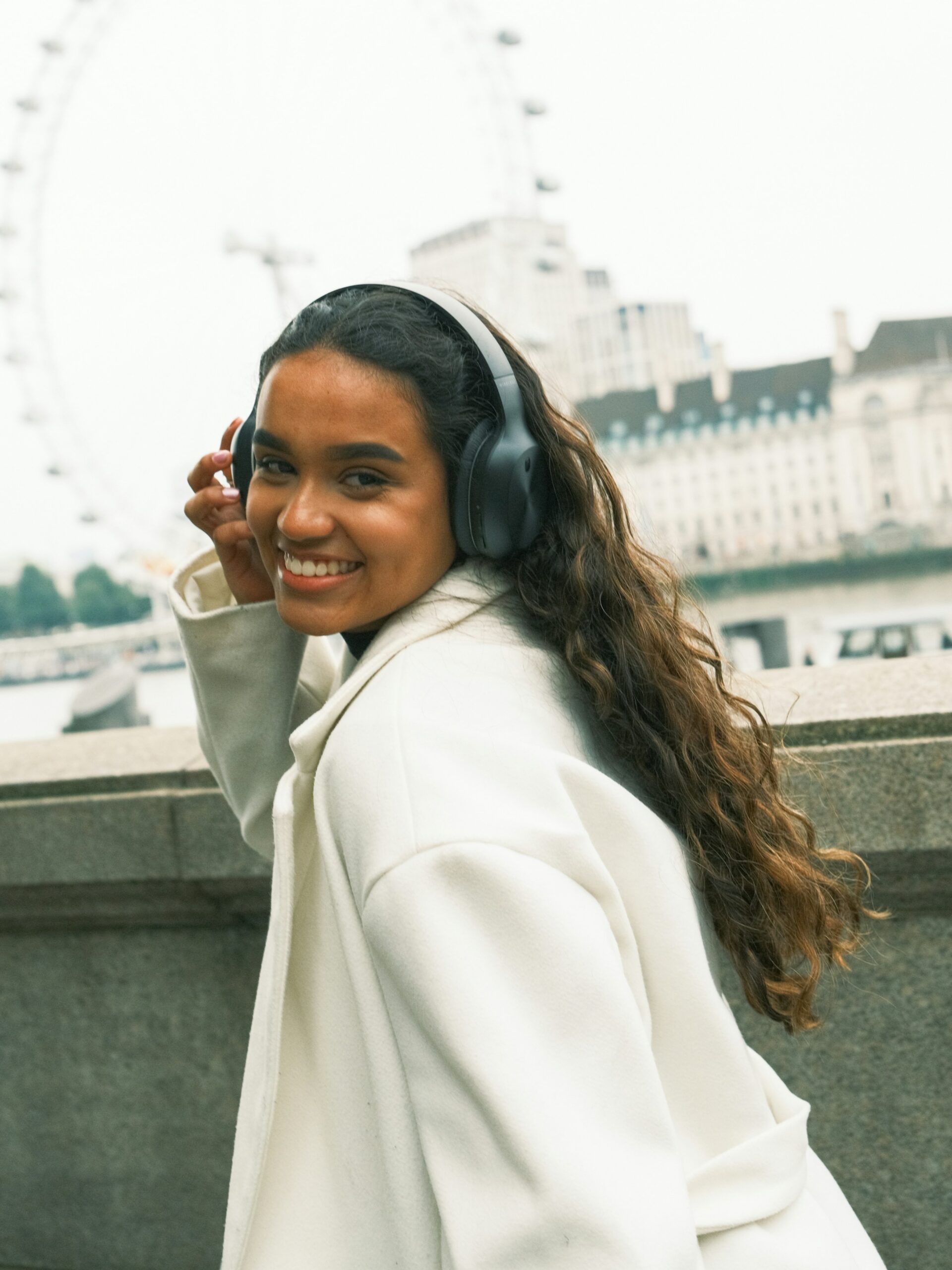 Une femme souriante avec un microblading des sourcils, portant un casque audio noir et un manteau blanc, se promenant en ville