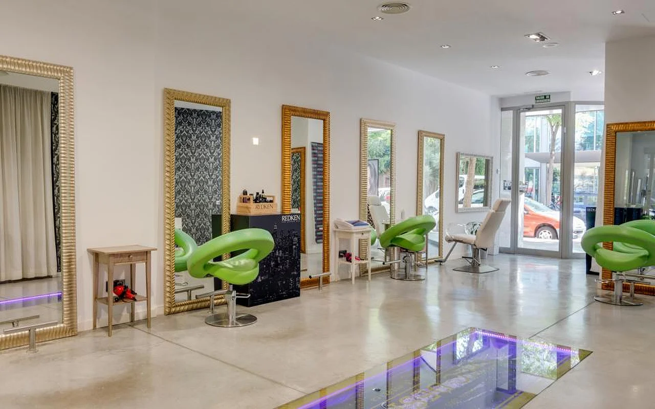 Un salon de coiffure lumineux et spacieux aux sols en béton ciré, de grands miroirs encadrés d'or le long d'un mur blanc, des fauteuils moulés vert citron, et une section au sol vitré éclairée d'une lumière violette, avec la rue visible à travers les portes vitrées au fond.