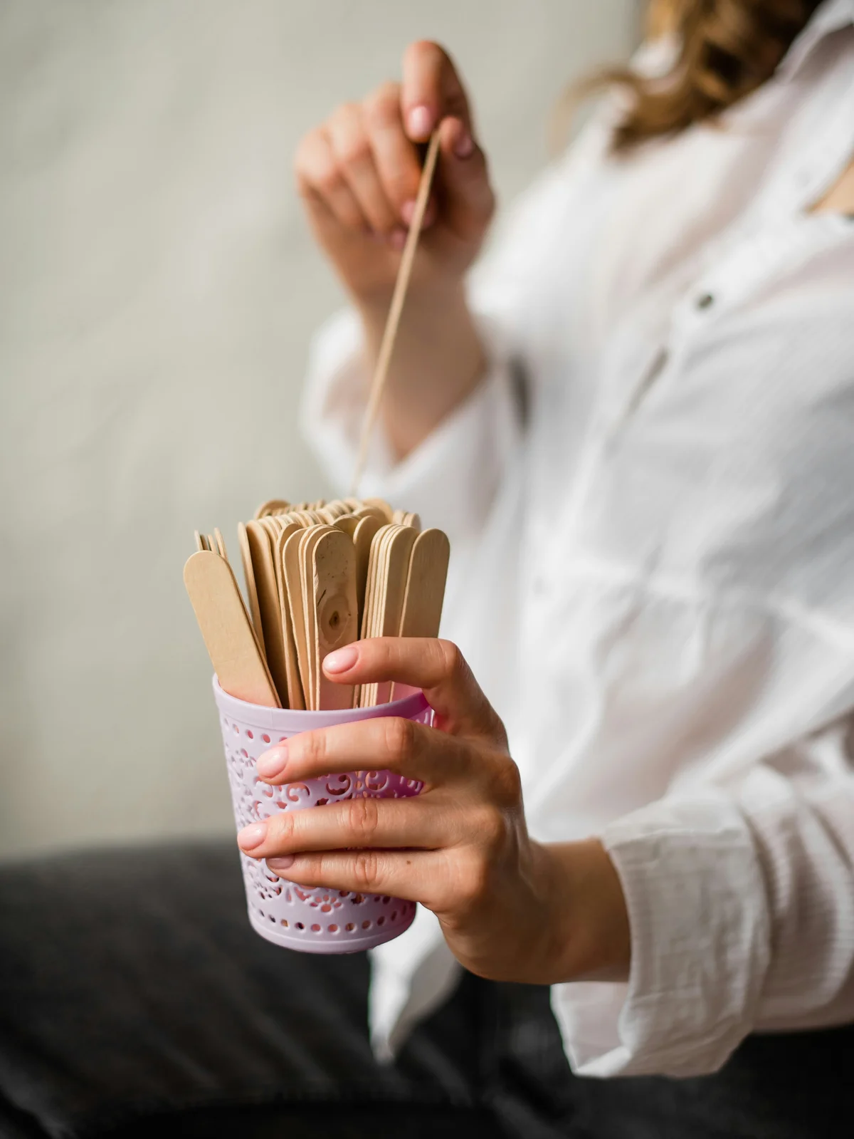 Gros plan sur une femme en chemise blanche tenant d'une main un pot rose rempli de spatules en bois et retirant une spatule de l'autre main