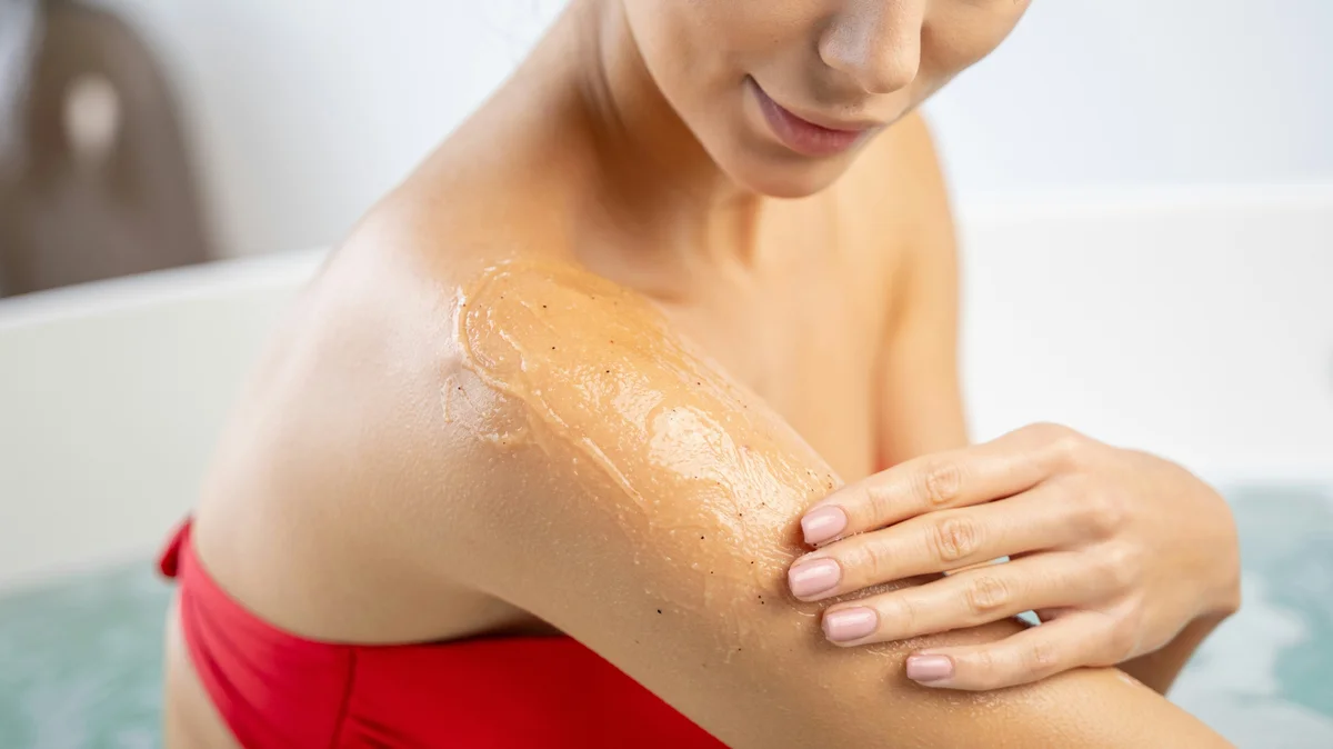Gros plan d'une femme en bikini rouge appliquant un gommage sur le haut de son bras dans une salle de bain claire et épurée