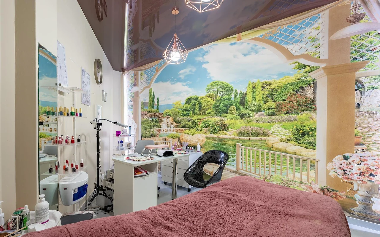 Une salle de soins beauté avec un lit drapé de rose et un poste de manucure blanc, devant une grande fresque murale de jardin et de lac, avec des étagères en verre garnies de produits à côté.