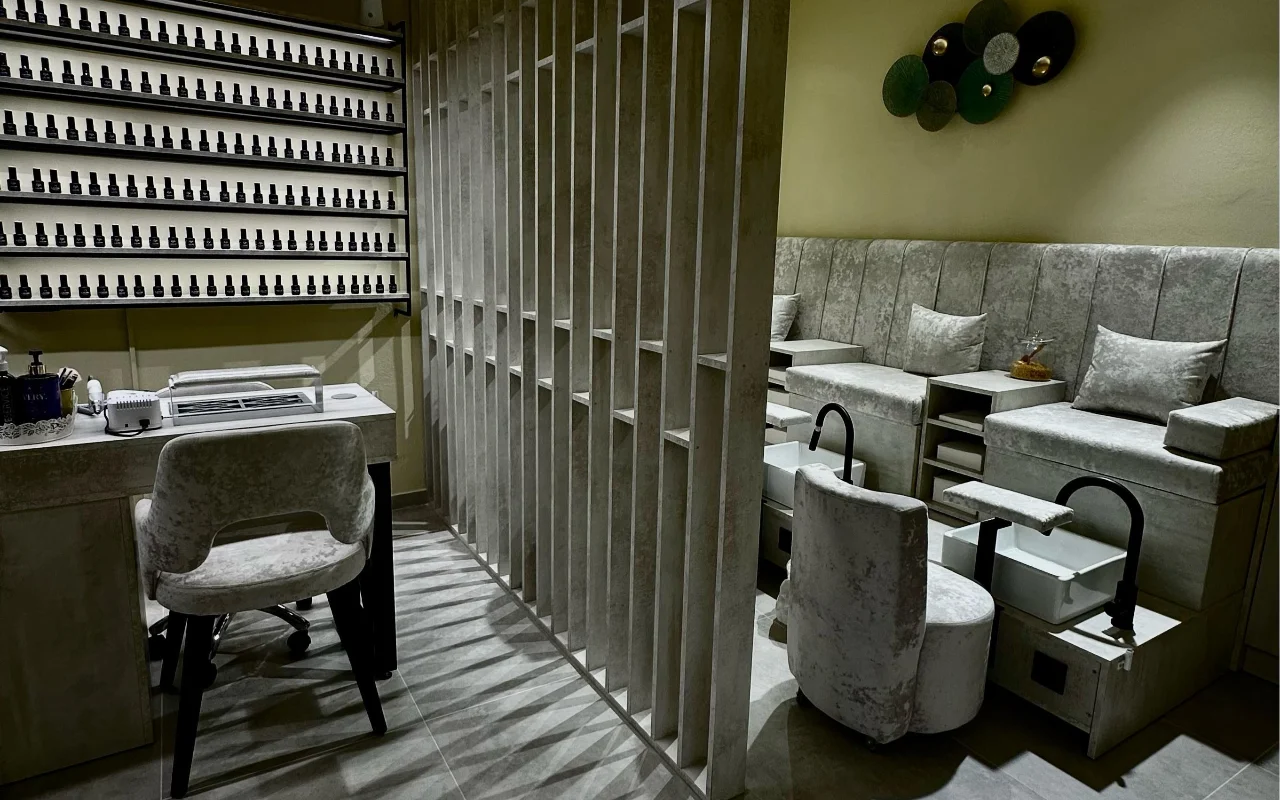 Une cabine de soin chez Sisu Nail Club and Academy à Périssa, Santorin, avec un mur entier de vernis à ongles, des fauteuils de manucure et pédicure en velours gris et un claustra en béton ajouré.
