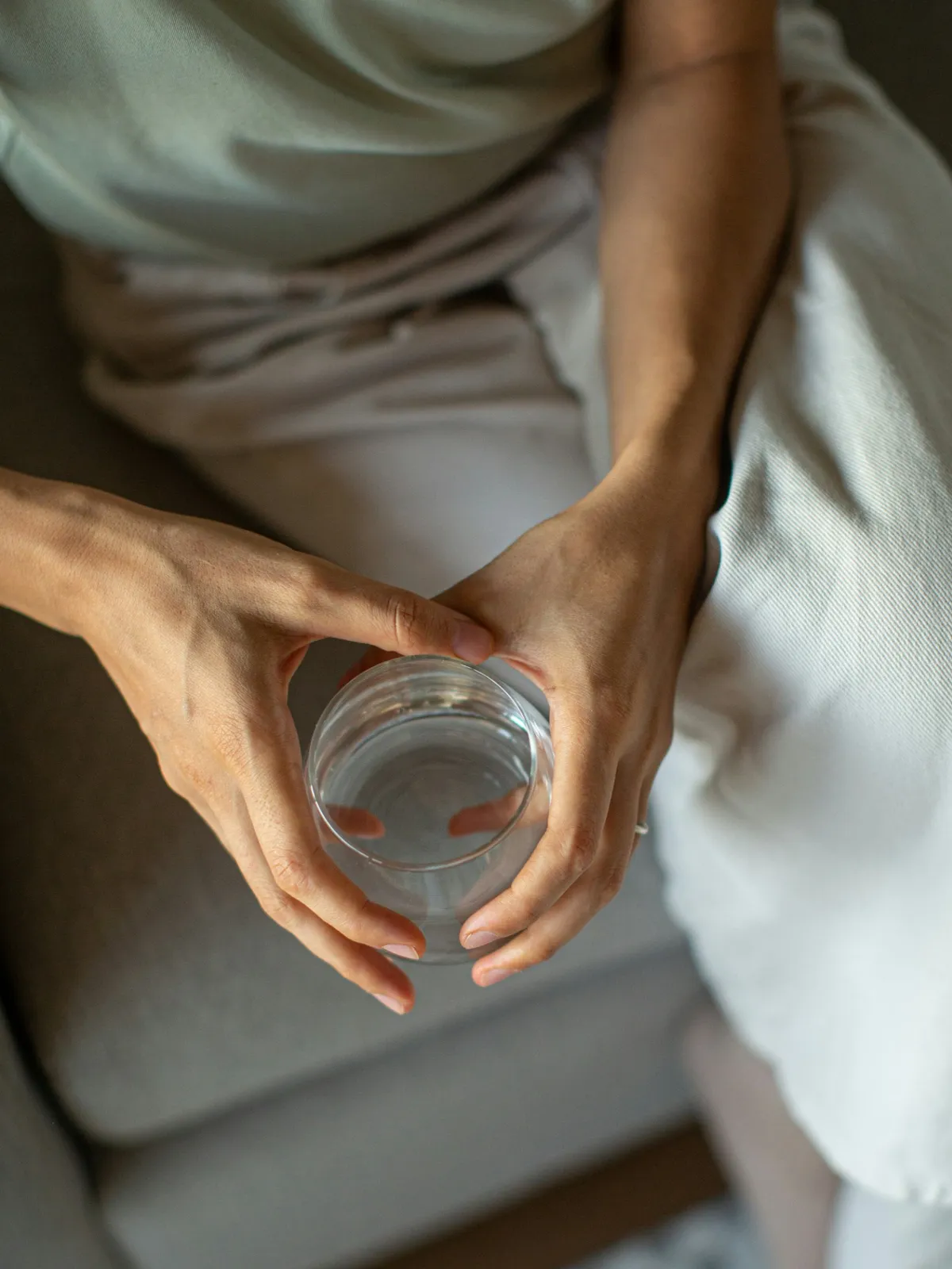 Vue de dessus d'une personne en vêtements amples assise sur un canapé gris, tenant un verre d'eau à deux mains
